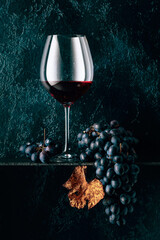 Glas Rotwein und blaue Trauben mit ausgetrockneten Weinblättern auf einem alten dunkelblauen Hintergrund. © Igor Normann