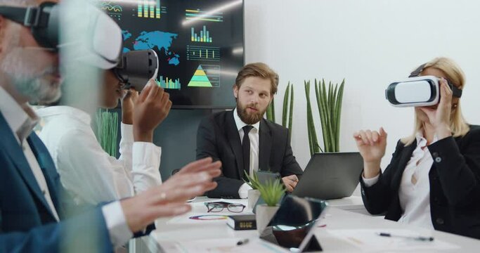 Attractive high-skilled international team working in virtual reality headset over joint project and talking with their bearded chief which entering datas on laptop