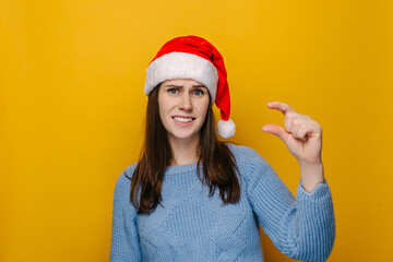 Young female shows very tiny object, shapes something small, wears Christmas hat and blue sweater, female looks in puzzlement, need some more, isolated over yellow studio wall. New Year concept