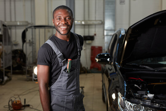 portrait of smiling african auto service worker at work place, young guy in uniform look at camera, during repairing a car