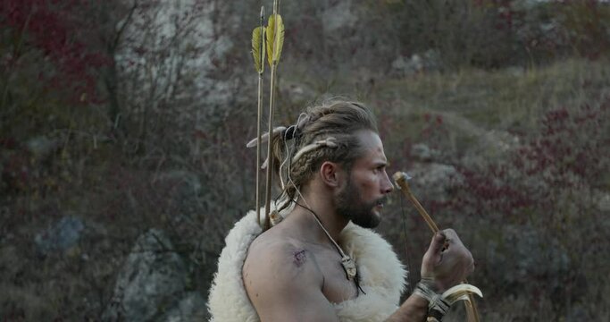 Wildman with bow and arrows hunting in nature. Side view of brutal bearded muscular savage male in fur vest taking arrow from quiver during hunting with bow in wild nature