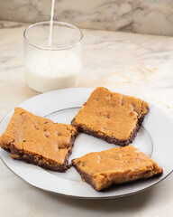 Blondies on white plate with chocolate chips falling on top. Decorated with milk, chocolate chips and Nutella. Delicious pastries out of the oven and ready to eat. Perfect for a bakery or online store