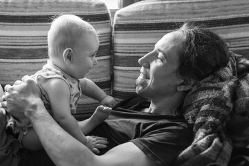 Handsome young man lying on couch and playing with infant baby boy by holding him sit on belly at home and spending time together
