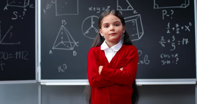 Portrait of cute funny Caucasian small teen clever girl with backpack standing in classroom at blackboard with math formulas, looking to camera and crossing hands. Smart pretty schoolgirl. Mathematics