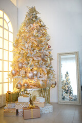 A Christmas tree with a Golden decoration is reflected in the mirror. New year, home decoration. Large window with yellow warm light, comfort in the living room, gift boxes