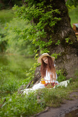 Cute red girl in straw hat on a flowering clearing at a picnic with fruit