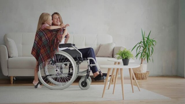 Candid handicapped woman in wheelchair using laptop computer at home, her daughter cover her with plaid and embrace mother, caregiving concept, 