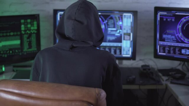 Back view of hacker in hood using multiple computers for cyber attack. Unrecognizable programmer coding or stealing information in Internet. Online security and global network.