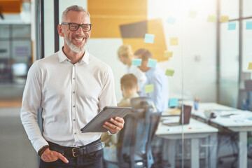 Happy mature businessman in classic wear holding digital tablet, looking at camera and smiling while standing in the modern office