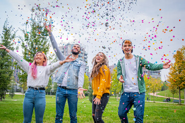 Group of stylish friends smiling and enjoying party throwing colorful confetti to the sky - Multietnic guys in the park throwing colorful confetti in the air