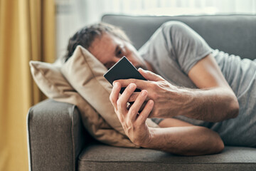 Depressed man using mobile phone while lying at living room sofa