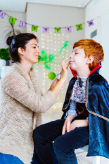 Vertical view of young mom doing vampire makeup to little kid at home for Halloween spooky costume party. Trick or treat for children and family. Seasonal and festive concept.
