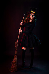 Young woman in black dress, witch hat and broomstick in hand posing on black background.