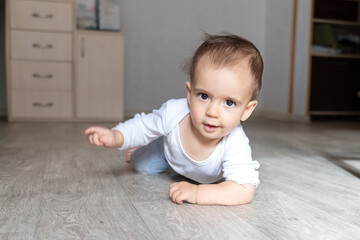 A little baby boy in white bodysuit at home is crawling . Beautiful portrait of a toddler. Big-eyed baby. 