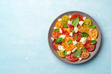 Italian caprese salad with varicolored tomatoes, mozzarella cheese, basil leaves, spices and olive oil in plate on concrete background