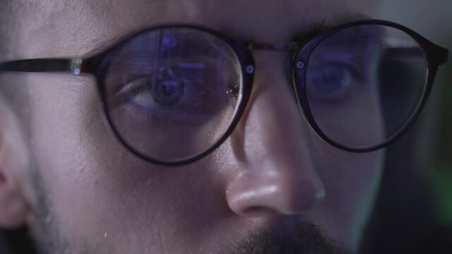Close-up eyes of Caucasian man with reflection of multiple computers screens on eyeglasses. Young concentrated hacker or programmer working online in global network.