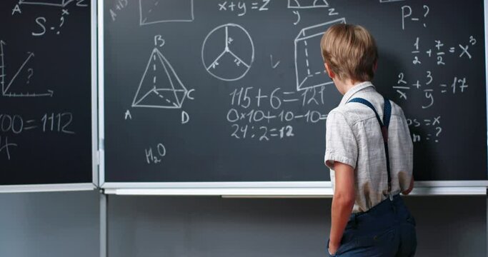 Rear on Caucasian small teen boy standing at blackboard in classroom and deciding math formulas and equations. Teenage smart schoolboy studying mathematics at chalkboard. Algebra or geometry.