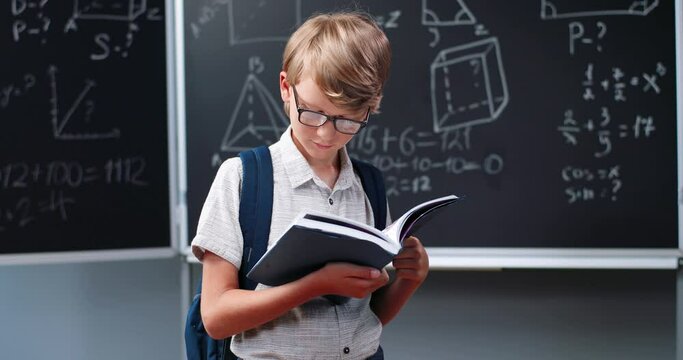 Caucasian teen boy in glasses with book in hands standing in school at blackboard with math formulas and studying. Teenager schoolboy holding and reading textbook. Mathematics.