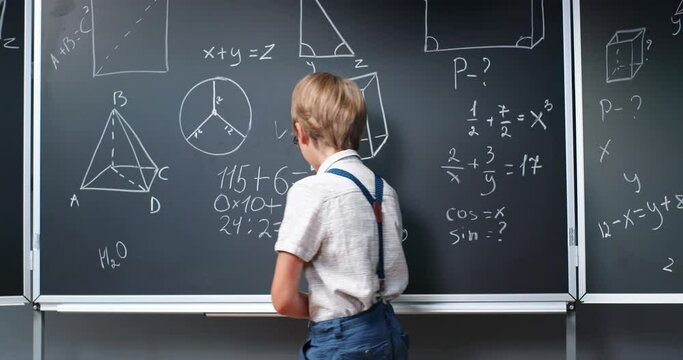Caucasian small boy in glasses thinking at blackboard in classroom, deciding math formulas equations and giving thumb up. Teenage smart schoolboy studying mathematics at chalkboard. Algebra. Geometry.