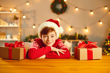 Fototapeta na wymiar Positive boy in Christmas clothing looking at camera at table surrounded by present boxes