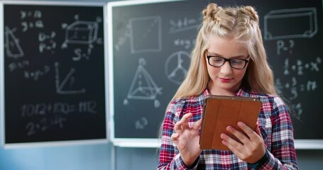 Joyful pretty Caucasian girl in glasses standing in school at blackboard with math and using tablet device. Teenage beautiful schoolgirl tapping and scrolling on gadget computer at mathematics class. - Powered by Adobe
