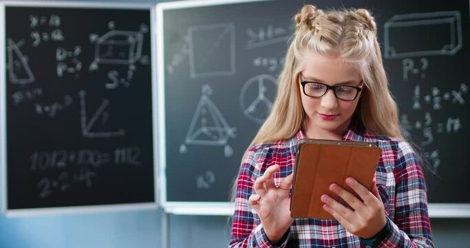 Joyful pretty Caucasian girl in glasses standing in school at blackboard with math and using tablet device. Teenage beautiful schoolgirl tapping and scrolling on gadget computer at mathematics class.