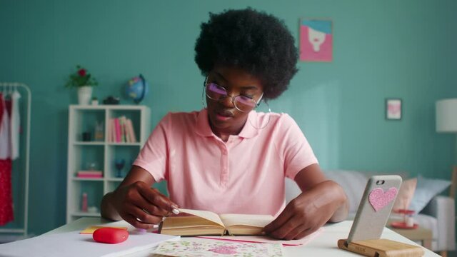 Young beautiful Afro-American woman, student, is studying at home, sitting at desk and reading interesting book, getting ready to literature class, foreground, Slow motion.