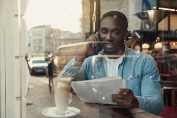 Handsome bearded dark-skinned businessman sitting near cafe window and talks on smartphone with business partner.