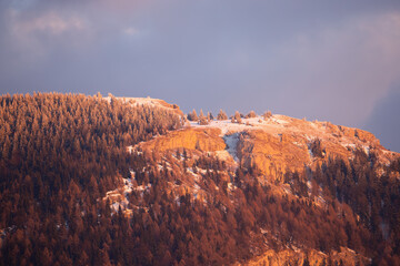 Winter nature scenery with a rocky mountainside covered with snow at sunset. Forest with trees growing on hill in wilderness. Horizontal telephoto view outdoor scene with sky and clouds.