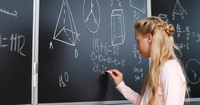 Caucasian small teen girl standing at blackboard in classroom and deciding math formulas and equations. Teenage smart schoolgirl studying mathematics at chalkboard. Algebra or geometry class.