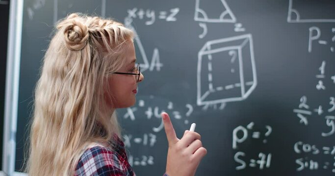 Caucasian small teen girl standing at blackboard in classroom, thinking and deciding math formulas and equations. Teenage smart schoolgirl studying mathematics at chalkboard. Algebra or geometry class