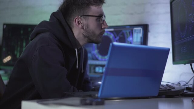 Concentrated programmer in eyeglasses working with multiple computers and laptop indoors. Portrait of absorbed young Caucasian man typing on keyboards and looking at screens.