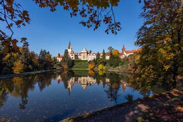 Pruhonice, Czech Republic - October 25 2020: Scenic view of famous romantic castle over a lake standing on green hill in park. Sunny autumn day with blue sky. Shapes of maple leaves in the foreground.