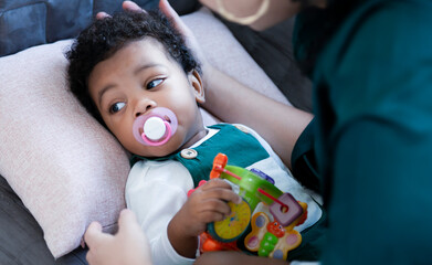 African American baby boy with pacifier is interesting his favorite toys while sit on mother's lap in living room. Portrait of a cute little happy black baby boy with mother at home, Family concept.