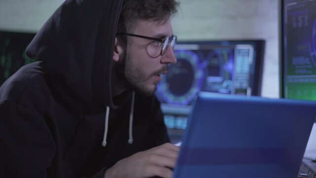 Portrait of focused young man hacking web site or launching computer virus using global network. Concentrated Caucasian programmer or hacker in eyeglasses and hood mining data in Internet.