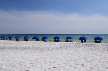 Vacationers on the beach sit under blue umbrellas and look into the ocean
