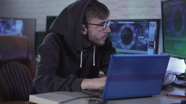 Serious bearded man in eyeglasses and hood typing on laptop keyboard and looking at computer screen. Young male Caucasian programmer hacking online site or password.