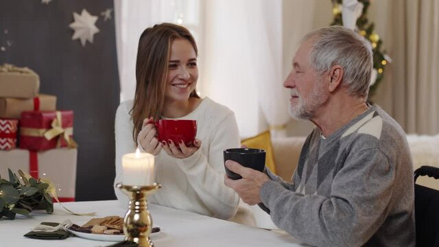 Young woman and senior grandfather in wheelchair indoors at home at Christmas, drinking tea.