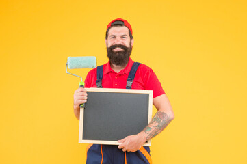hipster with copy space on board. repair and building. craftsman on construction. creativity. reconstruction advertising. bearded man with paint roller and blackboard. painter at painting works