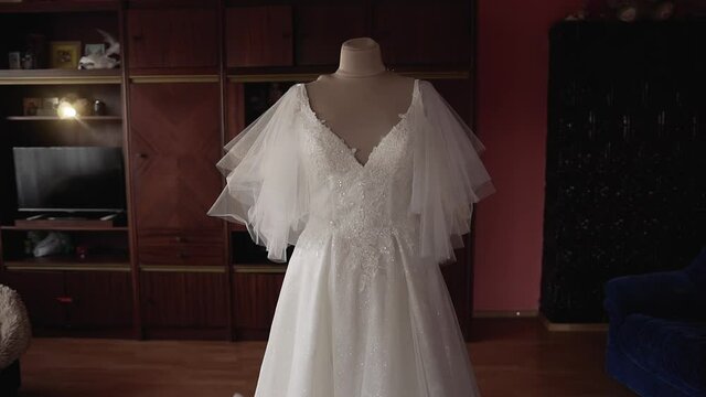 Wedding white lush beautiful dress with a train, sequins, lace and fatin on the shoulders stands on a beige mannequin in the middle of a dark room with a wooden wardrobe, sofa, two armchairs and oven