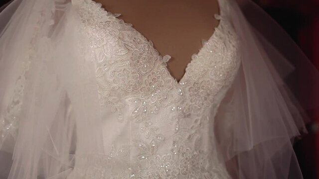 Bust of white lush wedding dress with lace and sequins on chest and belt with transparent fatin fabric that is sewn to a thin strap on the shoulders sits on a beige mannequin in a dark room. Close-up