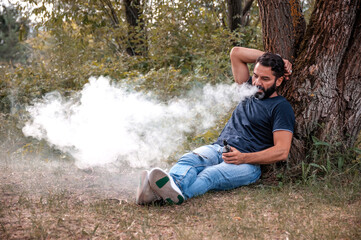 Naklejka na ściany i meble Stylish brutal smoker with beard and clouds of smoke looks relaxed. Hybrid cigarette.
