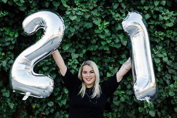 Smiling young woman holding number 21 balloons while standing against plants in park
