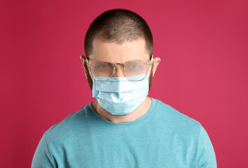 Man with foggy glasses caused by wearing disposable mask on pink background. Protective measure...