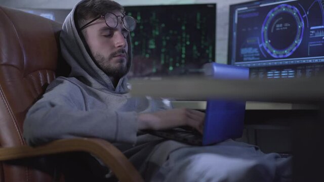 Exhausted Caucasian programmer sleeping on chair holding laptop. Computer monitors standing at the background. Portrait of young tired male hacker overworking at night.