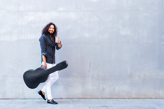 Smiling man with guitar showing thumps up while walking against gray wall