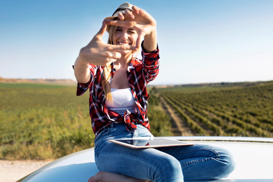 Smiling young woman showing finger frame while sitting on car roof