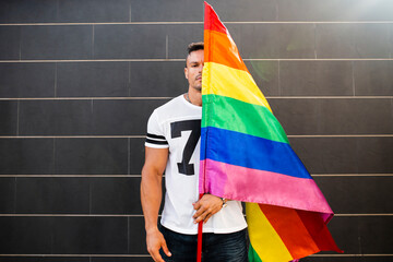 Young man holding rainbow flag while standing against wall