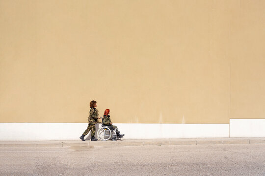 Female military office pushing wheelchair of army soldier while walking against wall on footpath