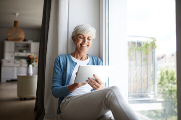 Smiling senior female using digital tablet while sitting on window at home